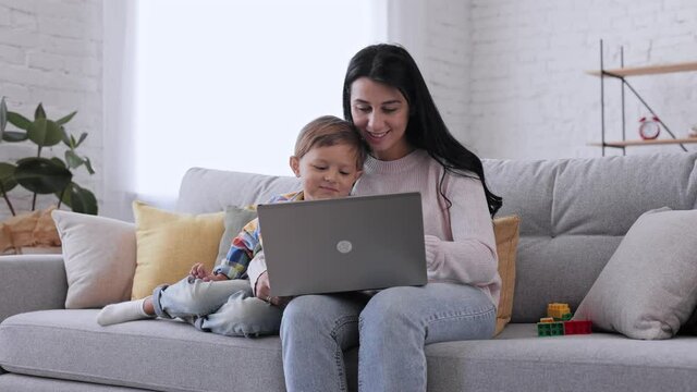 Young mother teaching little cute kid son use laptop showing website explaining device work, mother and small boy learning computer at home for child education. Family spend free time sit on sofa.