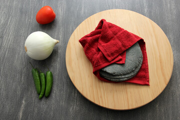 Fresh, delicious, freshly made blue dough tortillas along with a variety of fresh vegetables: onion, tomato, green chilies for Mexican sauce on a chopping board wrapped in red cloth
