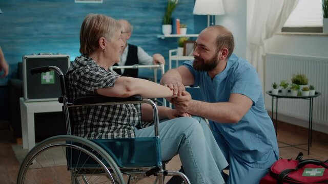 Man nurse comforting retired woman with disability in nursing home. Old person sitting in wheelchair and receiving medical checkup visit from healthcare specialist. Invalid patient