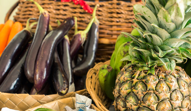 Pineapples, eggplants and carrots in rattan baskets on display at a small fruit and vegetable stand at an outdoor market.