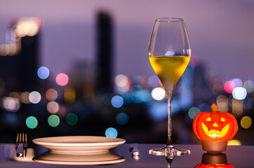 A glass of white wine with white plate, knife, fork and jack-o-lantern candle on city bokeh light...