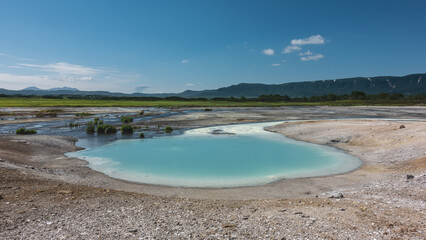 Obraz premium A small turquoise lake was formed from the hot springs. There are sulphurous deposits on the shores. A mountain range against a blue sky. Kamchatka. The caldera of the extinct volcano Uzon