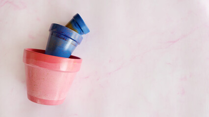 Flat lay of 3 terracotta pots in different sizes, from small to large. Pots are painted in blue and pink acrylic paints, and placed on pink background. With copy space on the right.