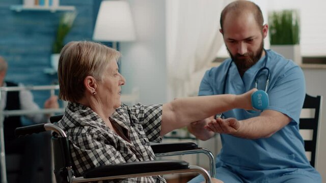 Patient with handicap doing physical exercises with dumbbells in nursing home. Man nurse giving assistance to senior woman in wheelchair with activity for recovery and healthcare.
