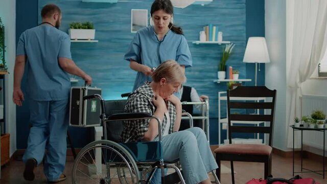 Nurse using stethoscope for heartbeat checkup on disabled woman in nursing home. Senior patient with disability in wheelchair receiving consultation from medical assistant for recovery