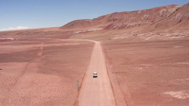 Aerial shot of white car driving along a red dirt road in the the Atacama Desert in Chile, South America near the salt lakes of Laguna Baltinache, red and orange mountains in the distance