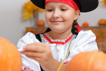 Celebrating Halloween at home. Close-up of a little girl dressed as a pirate. The child prepares for the party and draws on the pumpkin. - obrazy, fototapety, plakaty