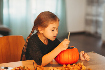 A little girl decorates a pumpkin in a room for Halloween. Kids decorate home. Children carve...