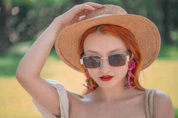 Beautiful redhead Asian Chinese girl wearing sunglasses, straw hat, knitted earrings. Bright sunny day, natural colors vivid background. Close up portrait of mature woman has fun in summer.