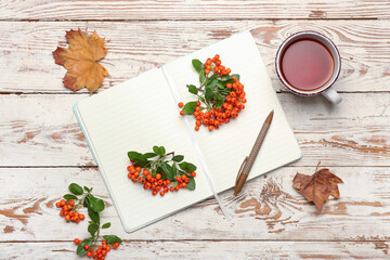 Composition with rowan berries, cup of tea, notebook and pen on light wooden background