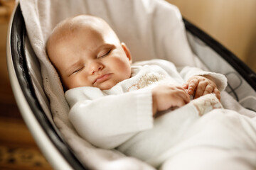 Cute baby laying in bouncer chair and sleep. Child relaxing in a swing. Adorable newborn baby in bodysuit in sunny bedroom. Family morning at home. Selective focus.