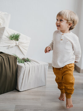 Two Years Old Caucasian Child Running Excited Around A Huge Pile Of Christmas Presents Wrapped In Furoshiki Technique And Adorned With Fir Tree Leaves