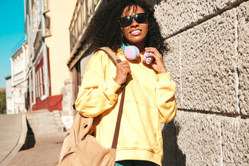 Beautiful black woman with afro curls hairstyle.Smiling model in yellow hoodie.Sexy carefree female enjoying listening music in wireless headphones.Posing on street background near wall in sunglasses