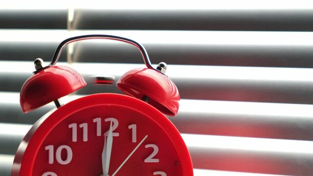 Red alarm clock rang at lunchtime at twelve clock, standing on windowsill against the background closed blinds.