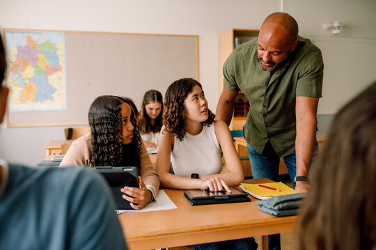 Male teacher teaching teenage girls over digital tablet at desk in classroom