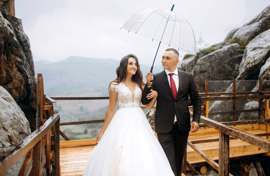 Bride and groom in mountainous terrain in the rain. Drops of rain. The bride and groom hold hands and look at the rain. Wedding photo shoot on the background of rocks.