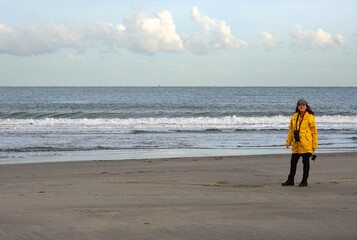 Lady with yellow raincoat on the beach. It's a windy day in autumn. The waves of the North sea are breaking on the shore