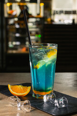 Glass of cold refreshing summer Blue Hawaiian cocktail made of  blue curacao liquor and slice of lemon and placed on slate board with ice cubes on counter in bar 
