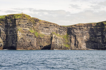 View on Cliff of Moher from a board of a ferry or cruise ship. West coast of Ireland. Popular tourist destination. National landmark. Cloudy sky. Calm surface of Atlantic ocean.