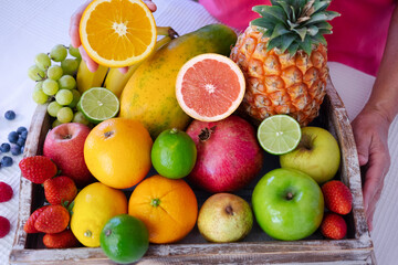 Wooden basket on white table full of fresh colorful fruits. Mature woman holding half orange fruit. Healthy eating and lifestyle