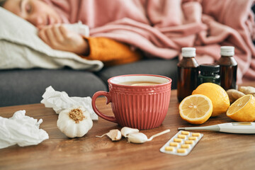 Hot tea, medicines and ill woman in the background lying on sofa