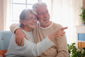 Smiling senior couple sitting at home hugging. Woman wearing eyeglasses indicating with a finger.  bright light from window