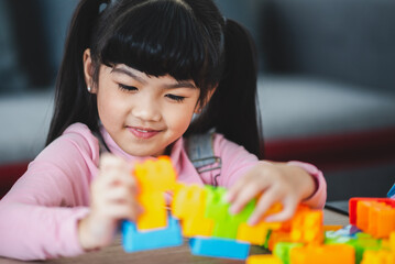 Cute little Asian girl playing alone with lego game blocks on table trying to build a new object while relaxing and enjoying