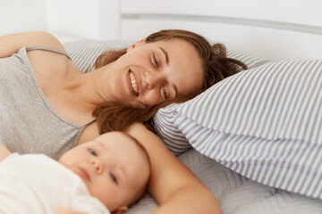Portrait of woman with infant baby girl or boy lying in bed in light room in early morning, enjoying weekend and spending time together, happy parenthood.
