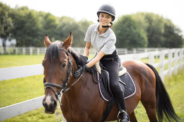 Female horseman riding brown Thoroughbred horse on green meadow near fence in countryside. Concept of rural resting and leisure. Idea of green tourism. Young smiling european woman. Sunny daytime