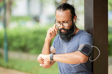 Serious young bearded Indian man in eyeglasses standing outdoors and adjusting earphones while listening to audio message on smartwatch