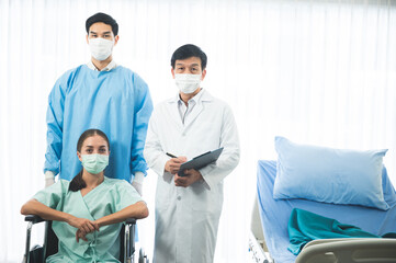 Portrait of two young male doctors standing wearing labcoat and surgeon uniform with female patient sitting on wheelchair with covid-19 face protective mask on all people in hospital