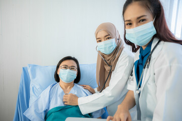 Portrait of two beautiful and cheerful female doctors wearing labcoat and stethoscope embracing senior patient sitting on hospital bed looking at camera