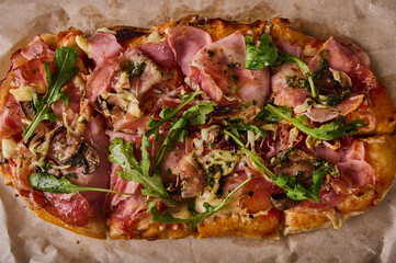 Traditional rectangular Roman pizza with prosciutto ham, tomatoes, mozzarella, mushrooms and arugula, top view, close up