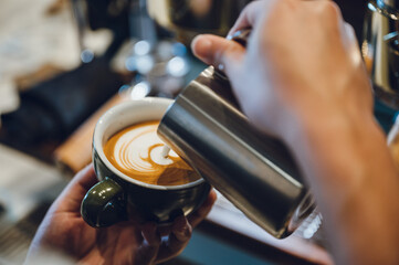 Closeup of hands of a young male barista pouring milk making latte art in a freshly brewed coffee in cafeteria counter for customer