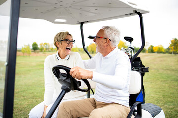 Portrait of healthy smiling senior couple driving golf car and heading to the green zone.