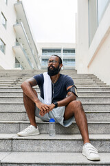 Pensive young Indian athlete with beard sitting on staircase and contemplating around