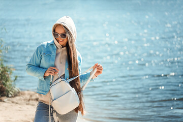 Portrait of a young stylish woman in a trendy casual outfit outdoors in a blue denim jacket with a leather white backpack in sunglasses.