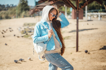 Portrait of a young stylish woman in a trendy casual outfit outdoors in a blue denim jacket with a leather white backpack in sunglasses.