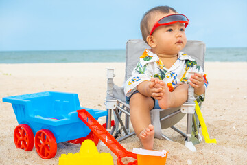 The 7-month-old baby was sitting in a chair on the beach with lots of toys.