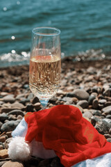A glass of champagne and a red Christmas hat on the shore of the blue sea.