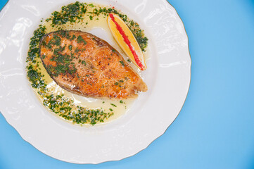 Grilled salmon steak with lemon and fresh herbs lies on a white plate on a blue background.