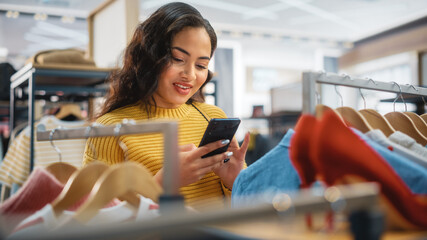 Beautiful Smiling Female Customer Shopping in Clothing Store, Using Smartphone, Browsing Online,...