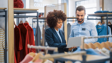 Clothing Store: Businesswoman Uses Laptop Computer, Talks to Visual Merchandising Specialist,...
