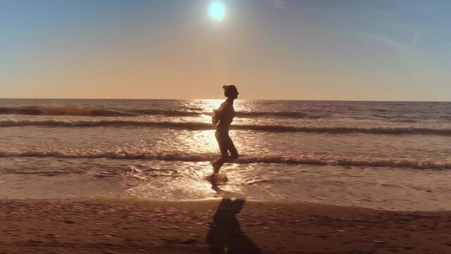 A slender woman runs and jumps on the waves of sea water on a sandy beach. on the surf. Splashes of water and foam on the background of the orange sun at sunset. Caucasian beauty is happy and joyful.