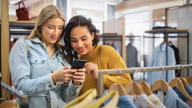 Two Beautiful Female Friends Shopping In Clothing Store, Using Smartphone, Browsing Online, Comparing On Internet. Customers Choosing Clothes. Fashionable Shop, Colorful Brands, Sustainable Designs.