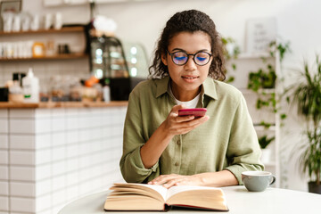 Young black woman using mobile phone while reading book at cafe