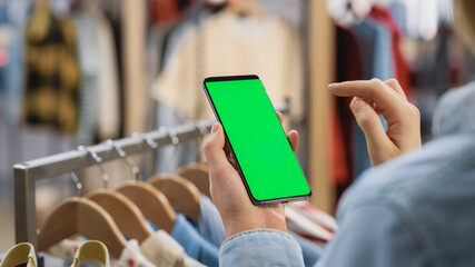 Clothing Store: Female Using Smartphone with Chroma Key Green Screen Display. Clothes Hanger with Stylish Branded Items for Retail Sale In the Background. Close Up Shot of a Mobile Device.