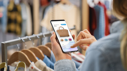 Clothing Store: Female Using Smartphone with Online Fashion Store User Interface to Chek Prices on a Stylish Branded Items. Close Up Over the Shoulder Shot of a Mobile Device.