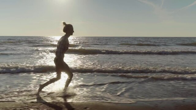 A slender woman runs and jumps on the waves of sea water on a sandy beach. on the surf. Splashes of water and foam on the background of the orange sun at sunset. Caucasian beauty is happy and joyful.