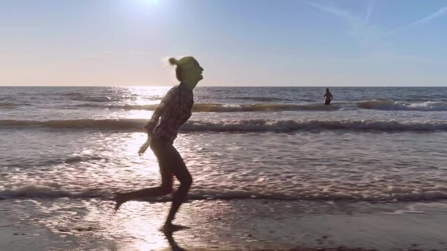 A slender woman runs and jumps on the waves of sea water on a sandy beach. on the surf. Splashes of water and foam on the background of the orange sun at sunset. Caucasian beauty is happy and joyful.